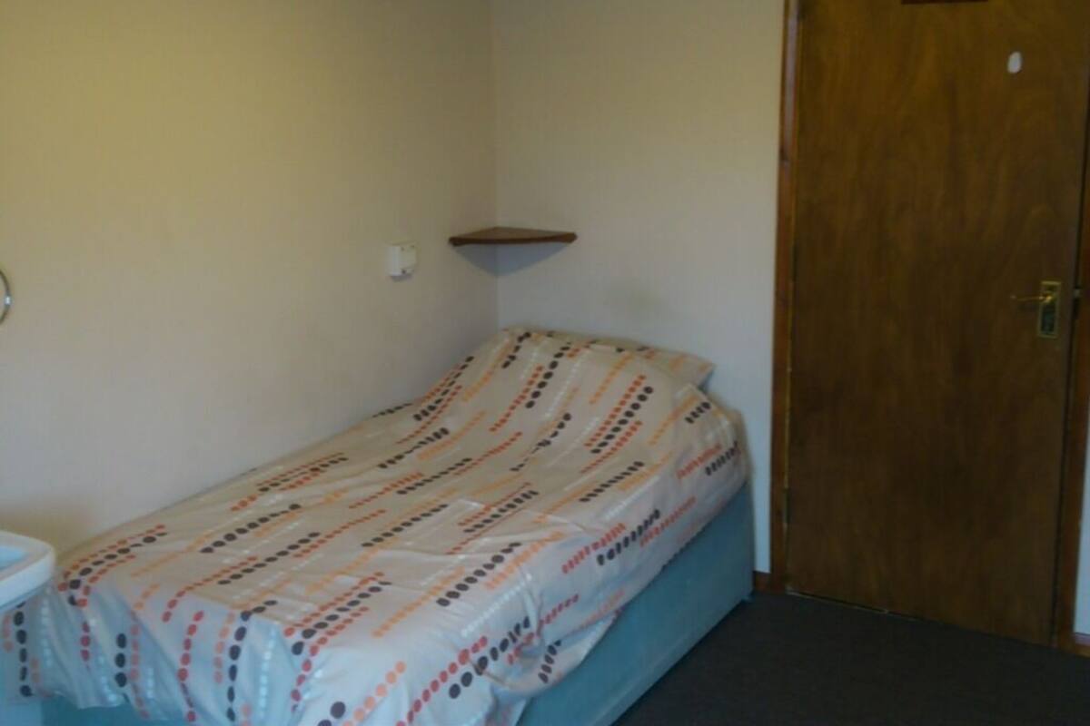 A small, single-bed room with a wooden door, a small shelf, and a bedside table.