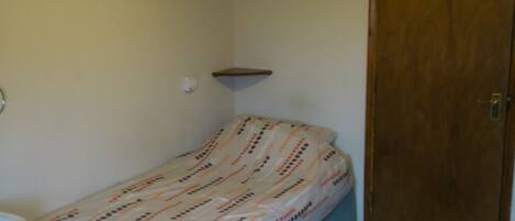A small, single-bed room with a wooden door, a small shelf, and a bedside table.