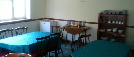 Un comedor con mesas y sillas, un reloj en la pared y una estantería con cuencos y tazas.
