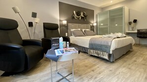 Una habitación de hotel moderna con una cama, dos sillones, una mesita con una revista, un televisor en la pared y un espejo grande.