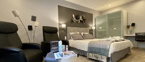 Una habitación de hotel moderna con una cama, dos sillones, una mesita con una revista, un televisor en la pared y un espejo grande.