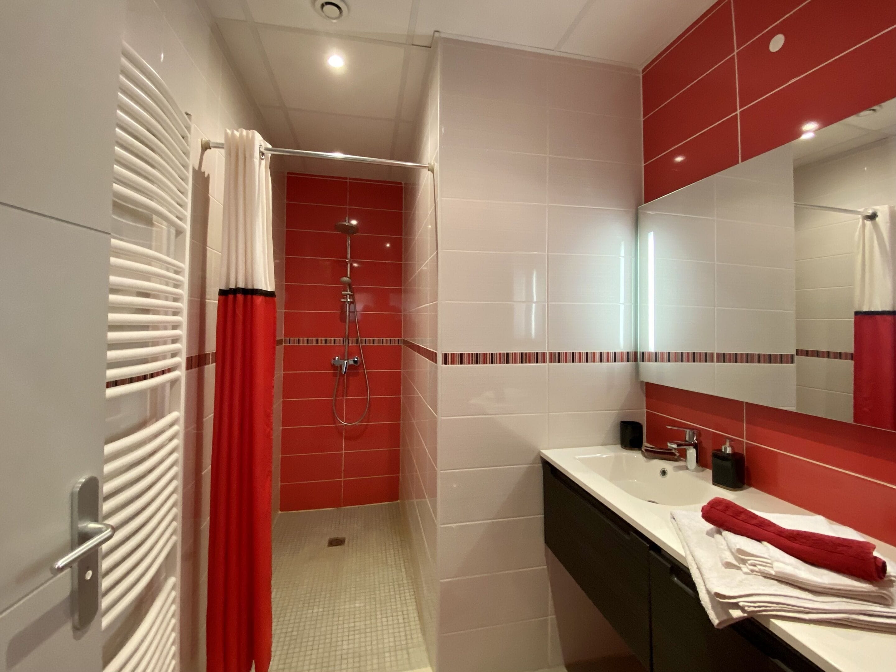 A modern bathroom with a red shower, white tiles, a large mirror, and a white sink with a red towel.