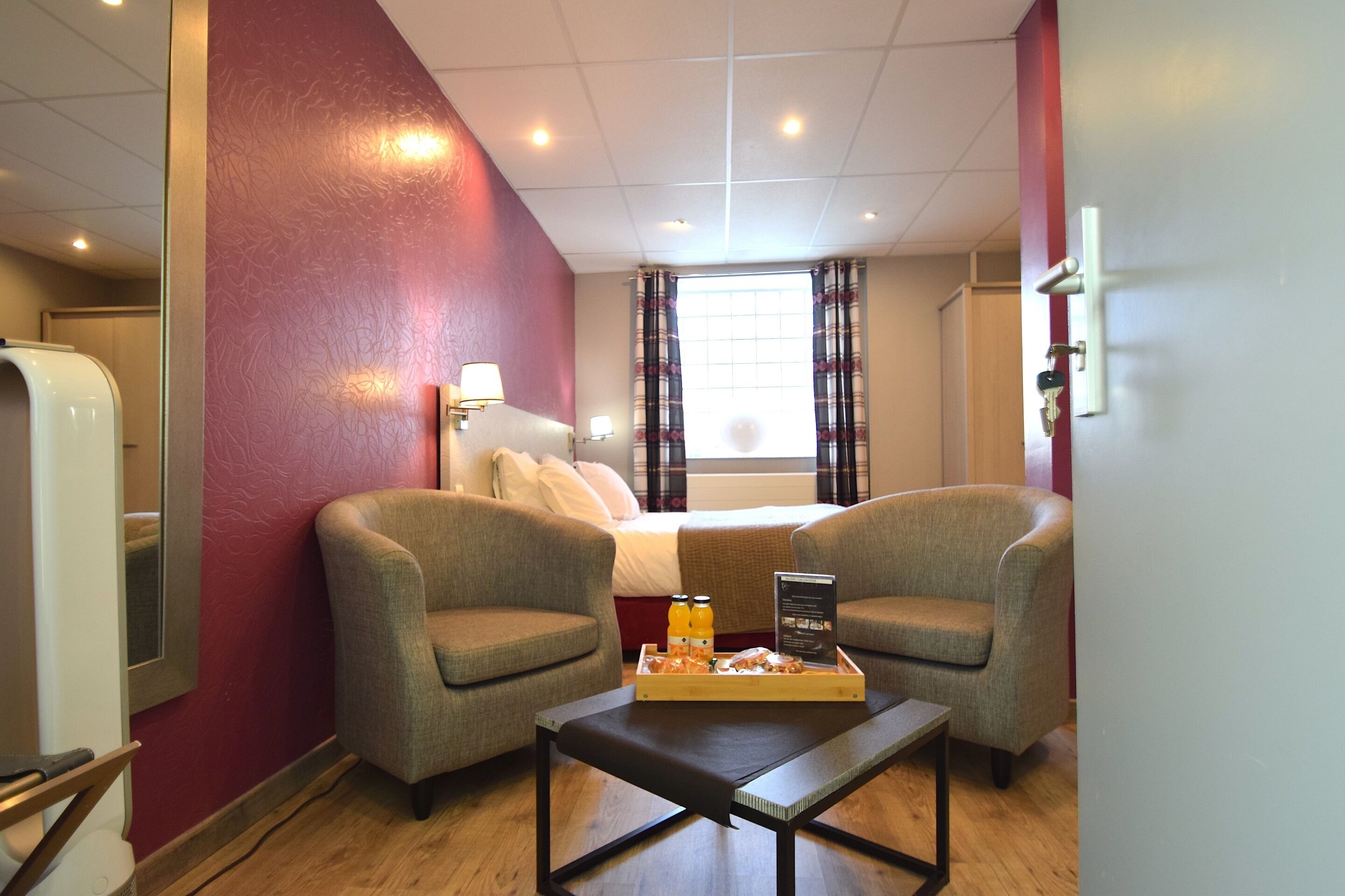 Une chambre d’hôtel comprenant un lit, deux fauteuils, une table basse avec des éléments pour le petit-déjeuner, un miroir et une fenêtre avec des rideaux.