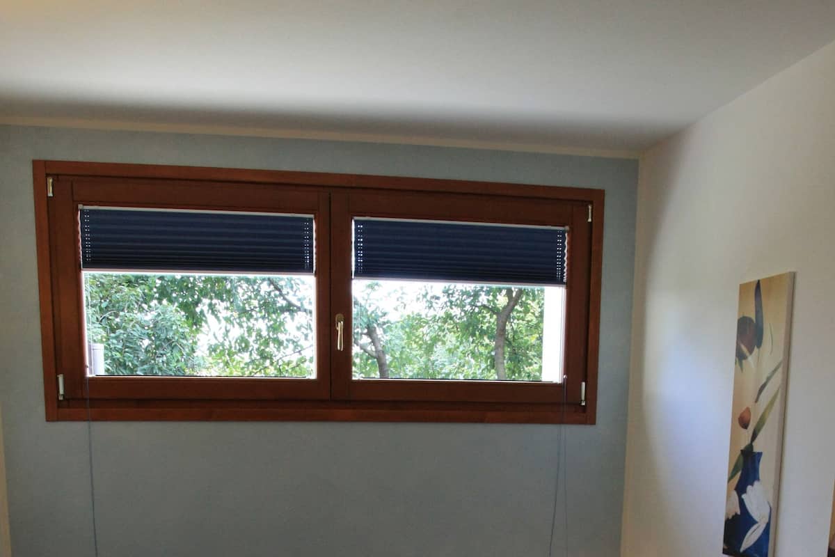Une chambre à coucher avec un lit, deux oreillers, une table de chevet, une lampe, une fenêtre avec des stores et un tableau au mur.