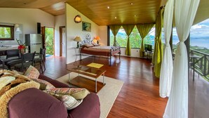 Habitación amplia con cama, escritorio y sofá. Se ve follaje a través de amplias ventanas.
