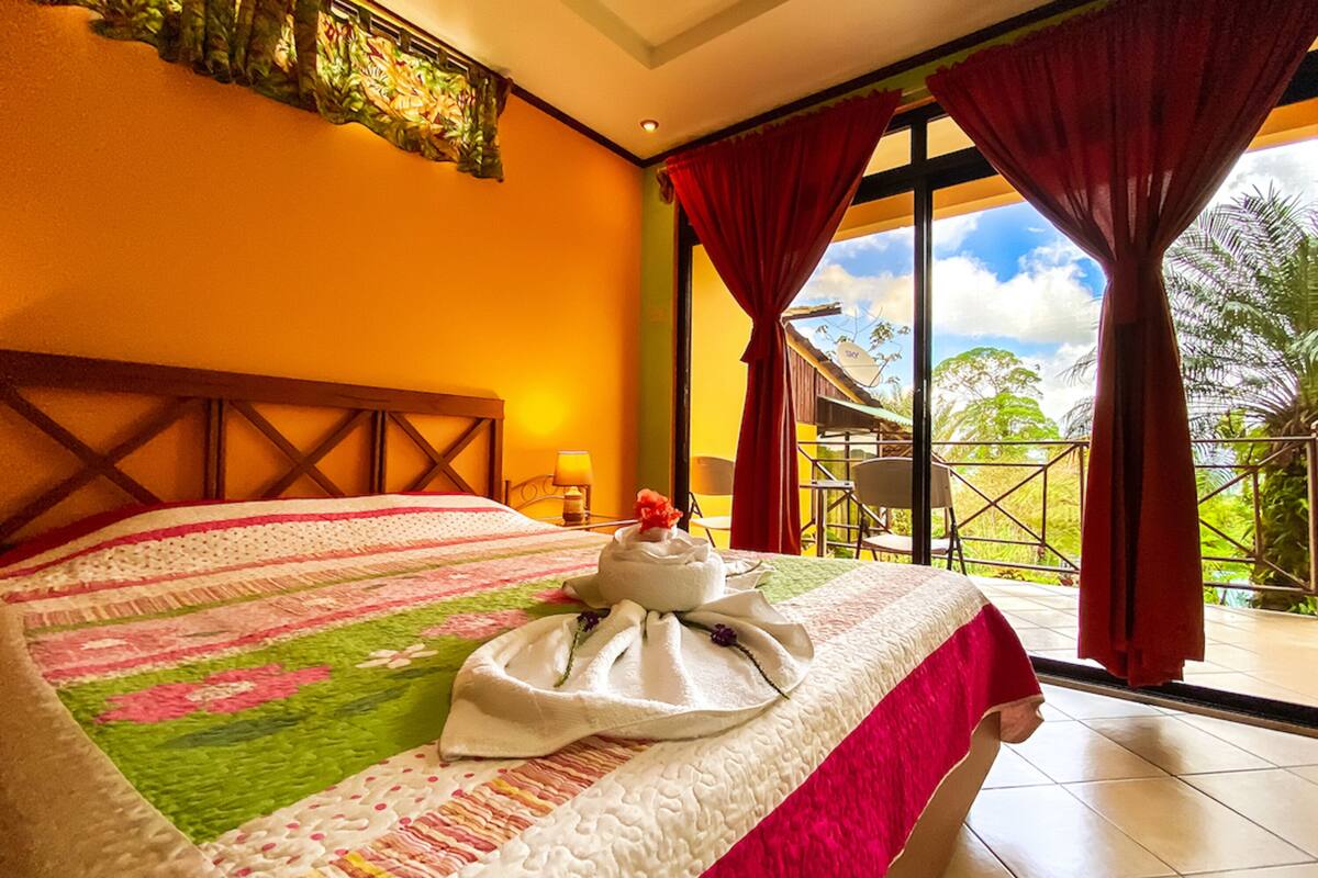 A hotel room with a large bed, a view of the outdoors, and a balcony with a table and chairs.
