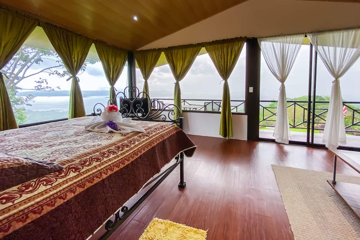 A bedroom with a wooden ceiling, a bed with a patterned bedspread, a view of the sea through large windows, and a balcony with a railing.