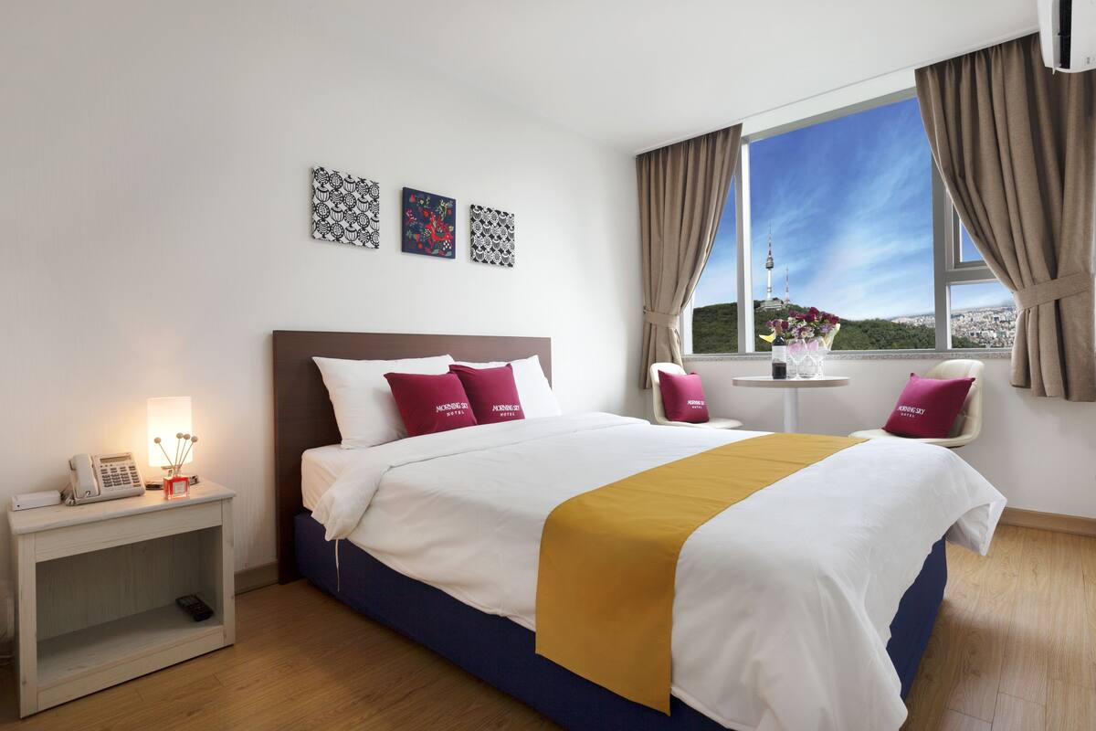 A hotel room with a large bed, a bedside table with a lamp, a small table with a vase of flowers, and a view of a tower through the window.