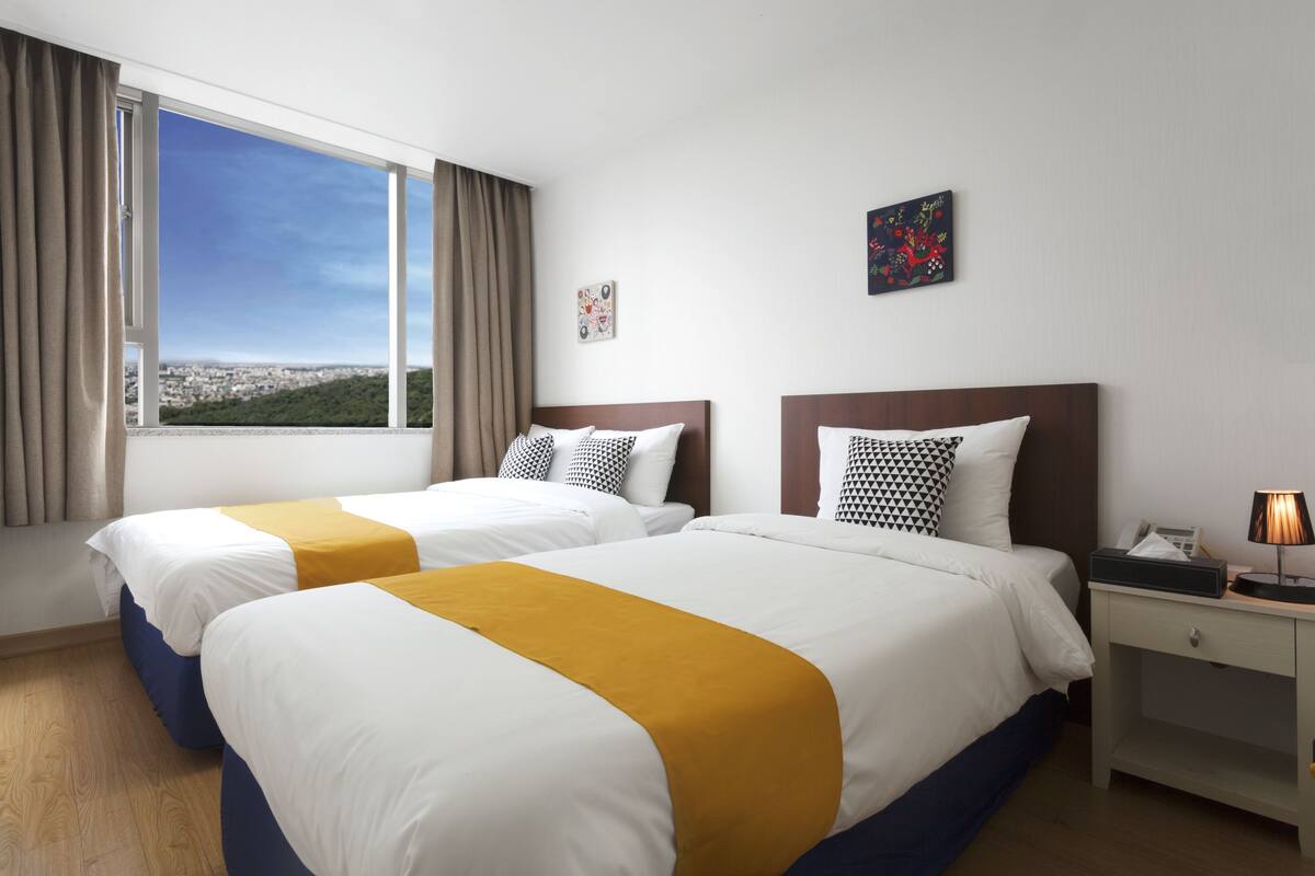A hotel room with two beds, a large window with a city view, a bedside table with a lamp, and artwork on the wall.