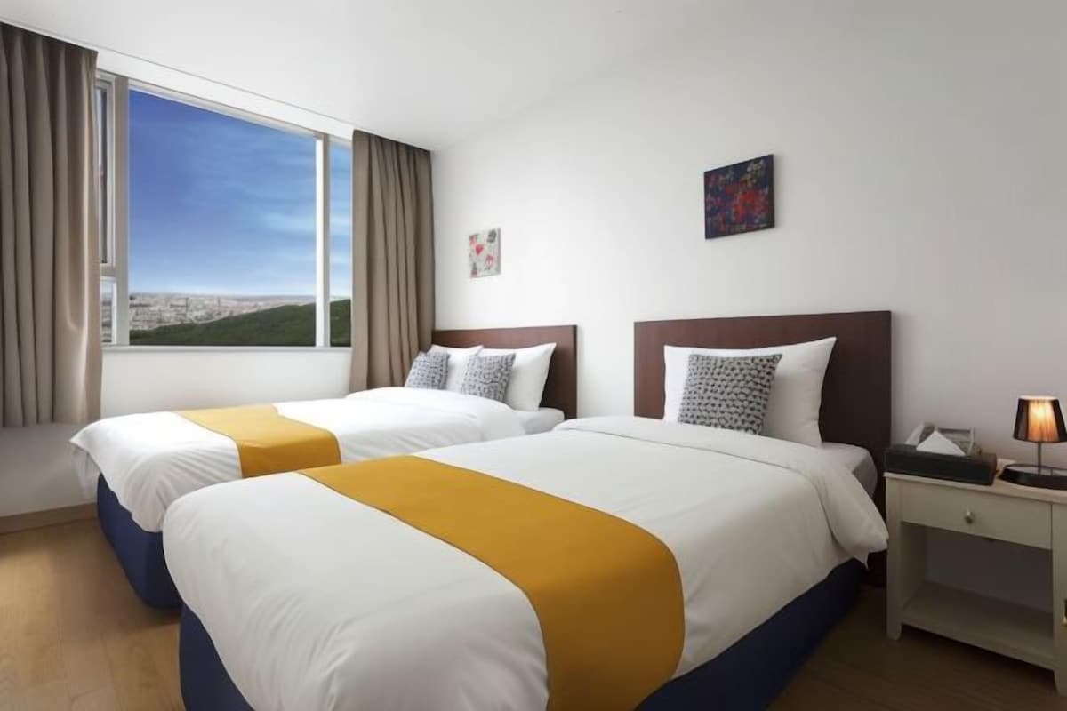 A hotel room with two beds, a large window with a city view, a bedside table with a lamp, and a small painting on the wall.