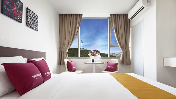 A hotel room with a bed, two chairs, a small table with a vase, and a view of a tower through the window. - Hotel Morning Sky (Seoul)