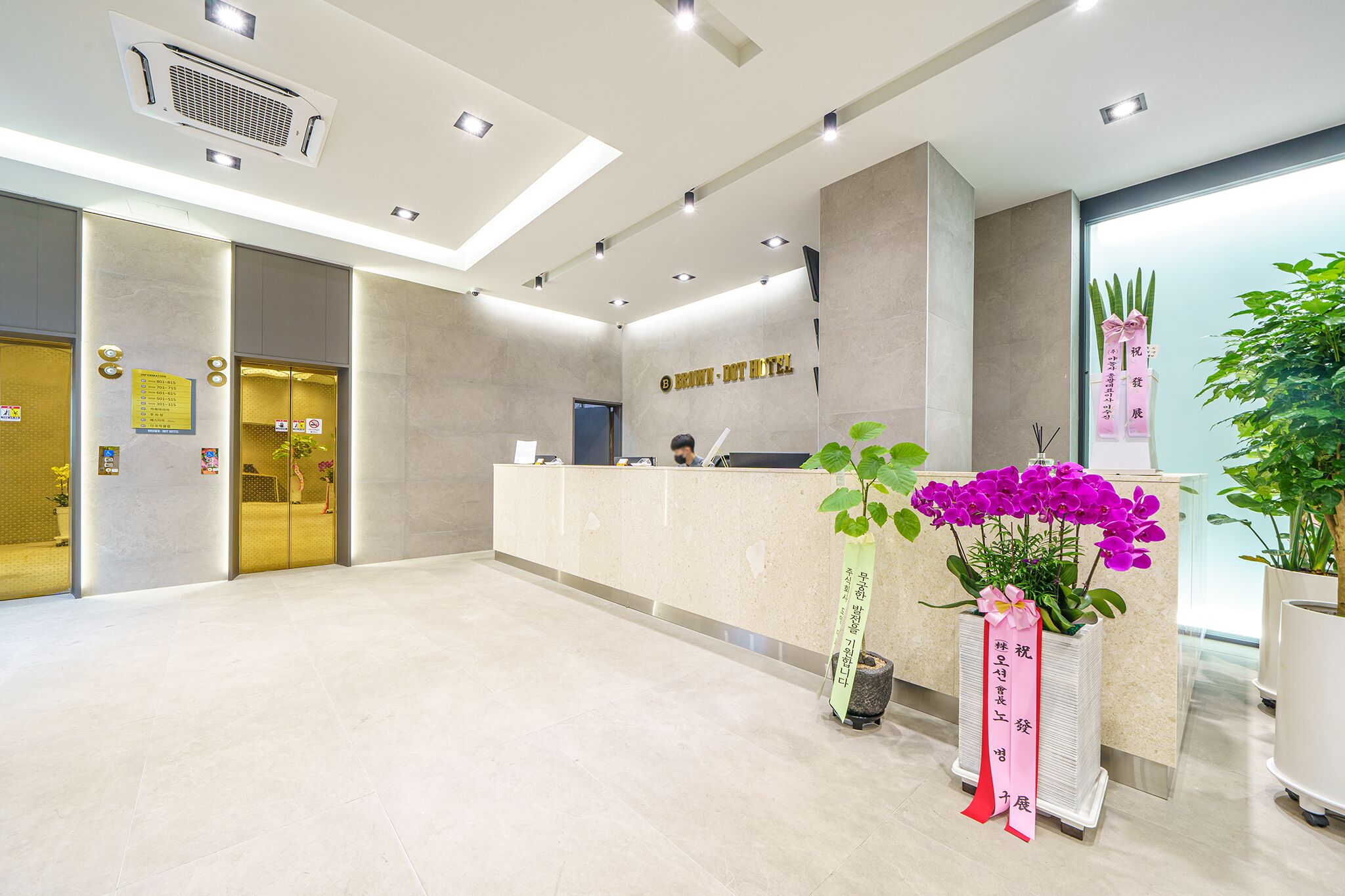 A hotel reception area with a modern design, including a reception desk, a digital screen, and a potted plant with purple flowers.