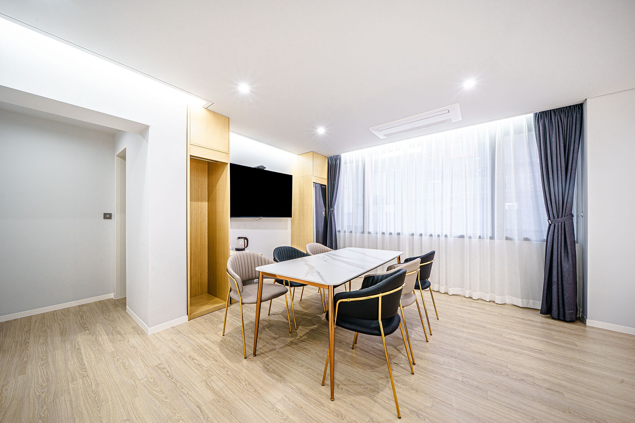 A modern, well-lit room with a wooden floor, a large table with chairs, a flat-screen TV, and a window with curtains.