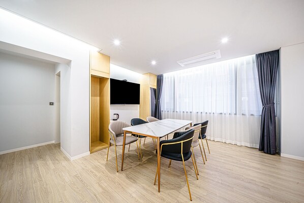 A modern, well-lit room with a wooden floor, a large table with chairs, a flat-screen TV, and a window with curtains.