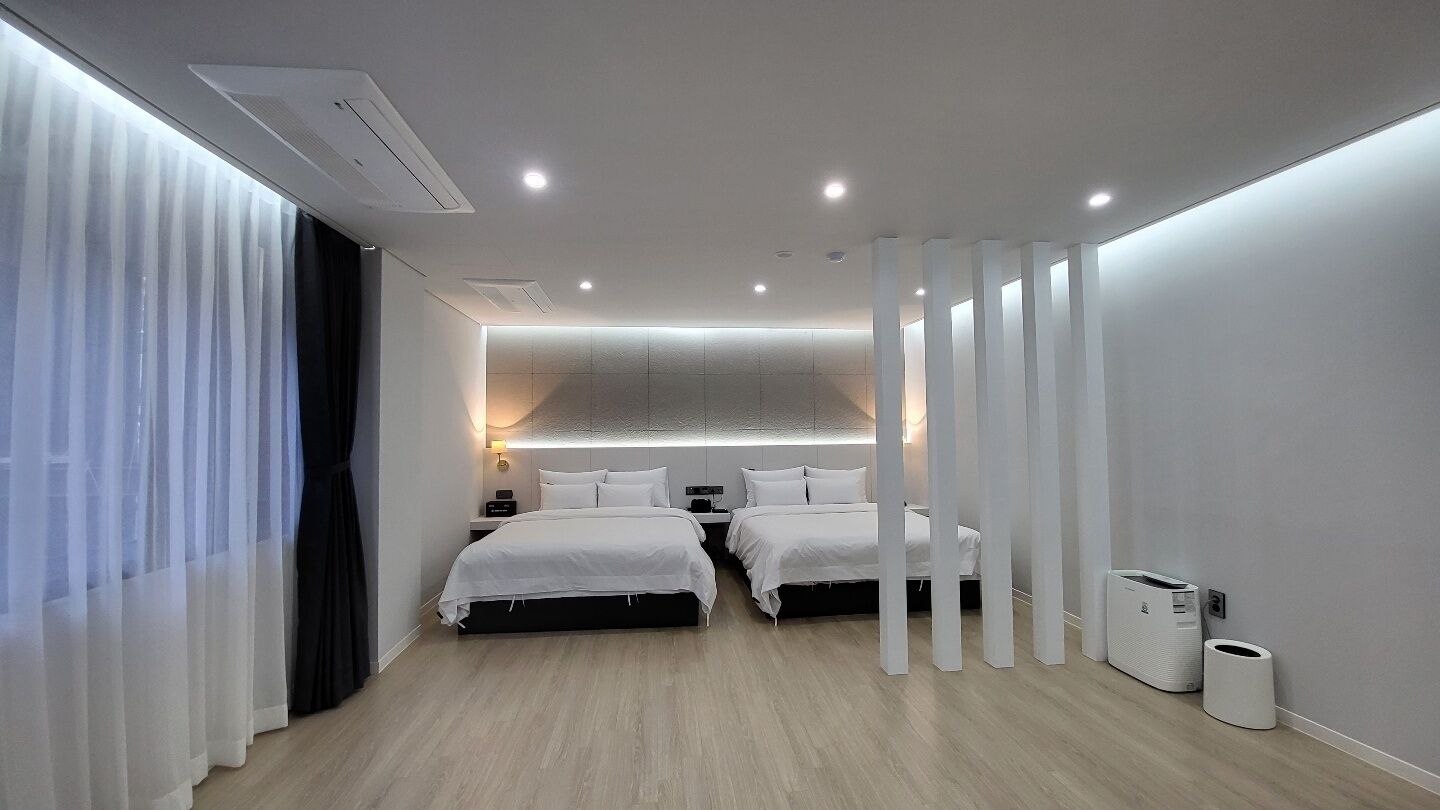 Una habitación de hotel moderna con dos camas, un ventanal con cortinas translúcidas, una lámpara empotrada en el techo y un cubo de basura blanco.