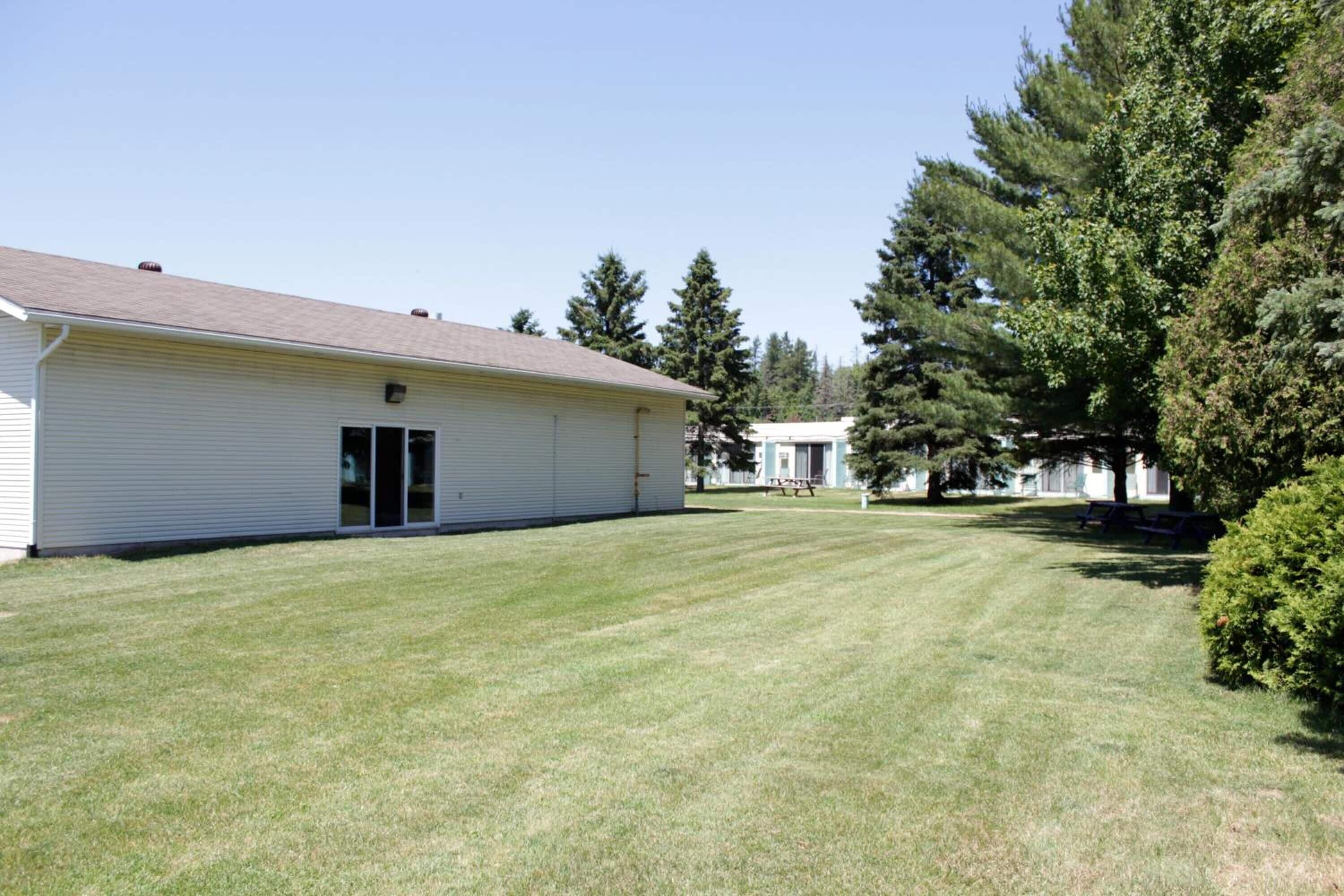 A single-story building with a flat roof, a large grassy yard, and a dense tree line in the background.