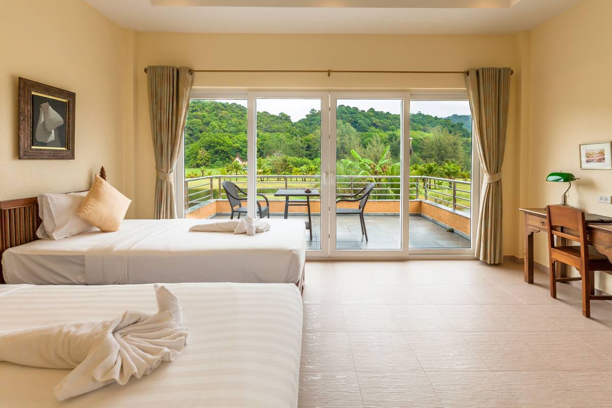 A hotel room with two beds, a desk, and a balcony with a view of the forest.