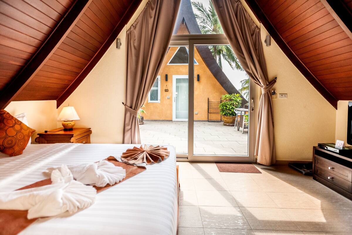 A hotel room with a bed, a nightstand, a lamp, a TV, and a sliding glass door leading to an outdoor area with a plant and a door.