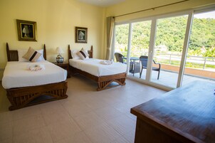Habitación de hotel con dos camas, un balcón con mesa y sillas, y amplias ventanas que dan a vistas del exterior.