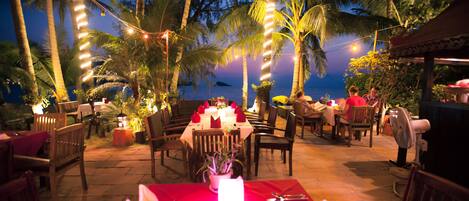 An outdoor dining area with tables set for two, surrounded by palm trees and a view of the ocean.