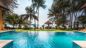 A swimming pool with clear blue water, surrounded by palm trees and a thatched-roof gazebo.