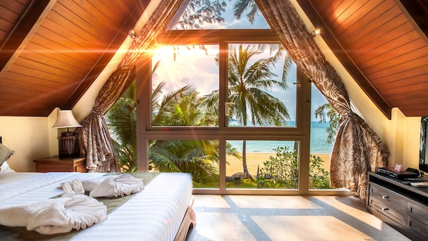 A bedroom with a large window overlooking a beach, a bed with towels, and a nightstand with a lamp.