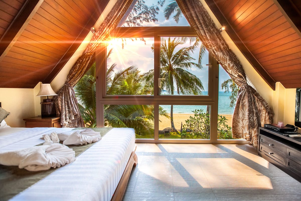 A bedroom with a large window overlooking a beach, a bed with towels, and a nightstand with a lamp.