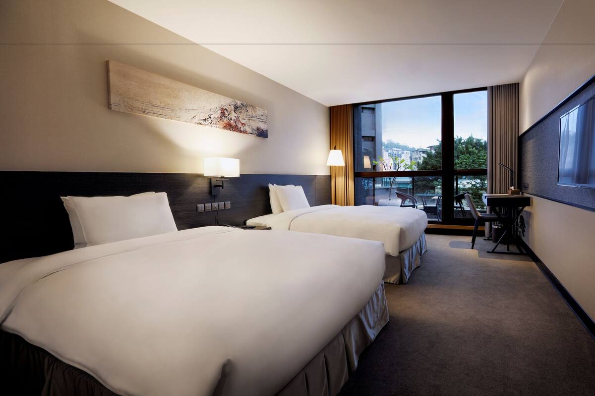 Een hotelkamer met twee bedden, een groot raam, een bureau en een schilderij aan de muur.