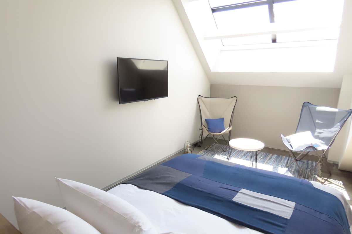 Une chambre d’hôtel avec un lit, un téléviseur fixé au mur, deux chaises, une petite table et une lucarne.