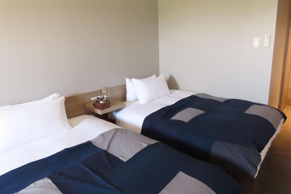 Ein Hotelzimmer mit zwei Betten, beide mit weißer und dunkelblauer Bettwäsche. An einem Nachttisch steht eine Lampe und eine kleine Schachtel.