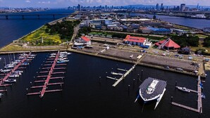 Une vue aérienne d’une marina où sont amarrés des bateaux, avec un pont en arrière-plan et le profil d’une ville qui se dessine au loin, de l’autre côté de l’eau.
