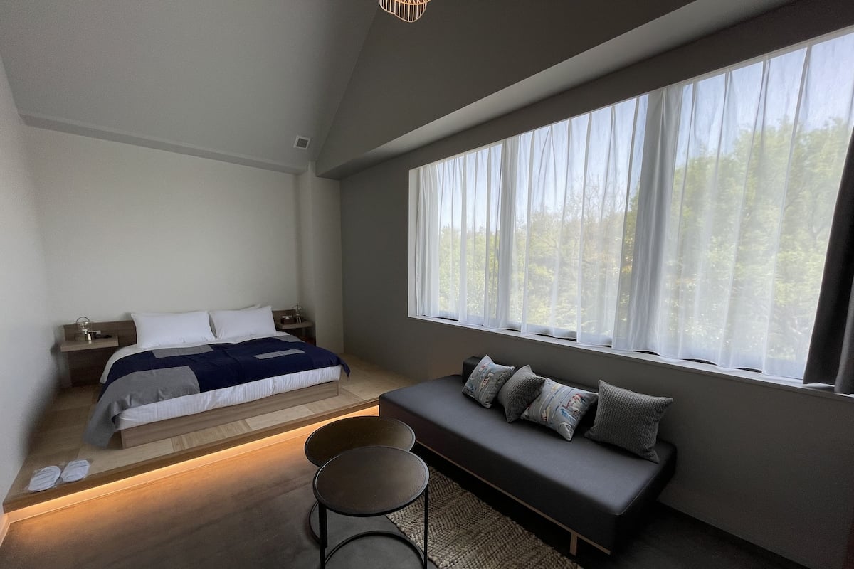Une chambre à coucher moderne avec un lit, des tables de chevet, un banc et de grandes fenêtres.