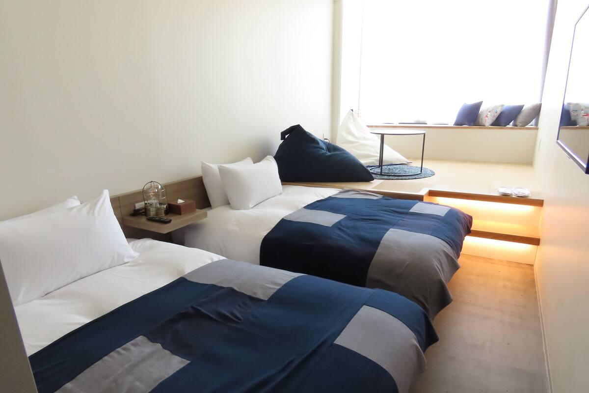 Ein Hotelzimmer mit zwei Betten, einem Nachttisch mit Lampe, einem kleinen Tisch mit Stuhl und einem Fenster mit Ausblick.