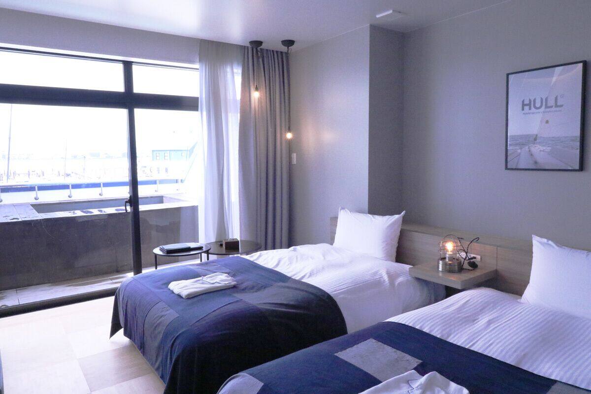 Une chambre d’hôtel avec deux lits, une grande fenêtre avec des rideaux, une petite table avec une lampe et un tableau encadré au mur.