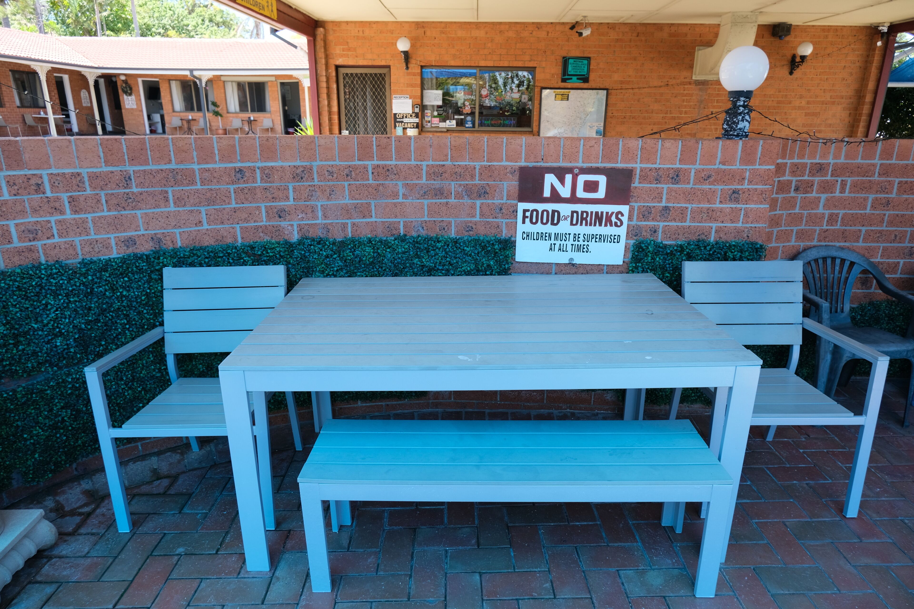 Een terras met een tafel en bankjes, een bakstenen muur en een bordje met de tekst 'GEEN ETEN EN DRINKEN'.