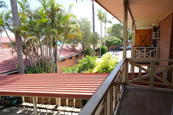 A balcony with a red roof overlooking a residential area with palm trees and a brick building.