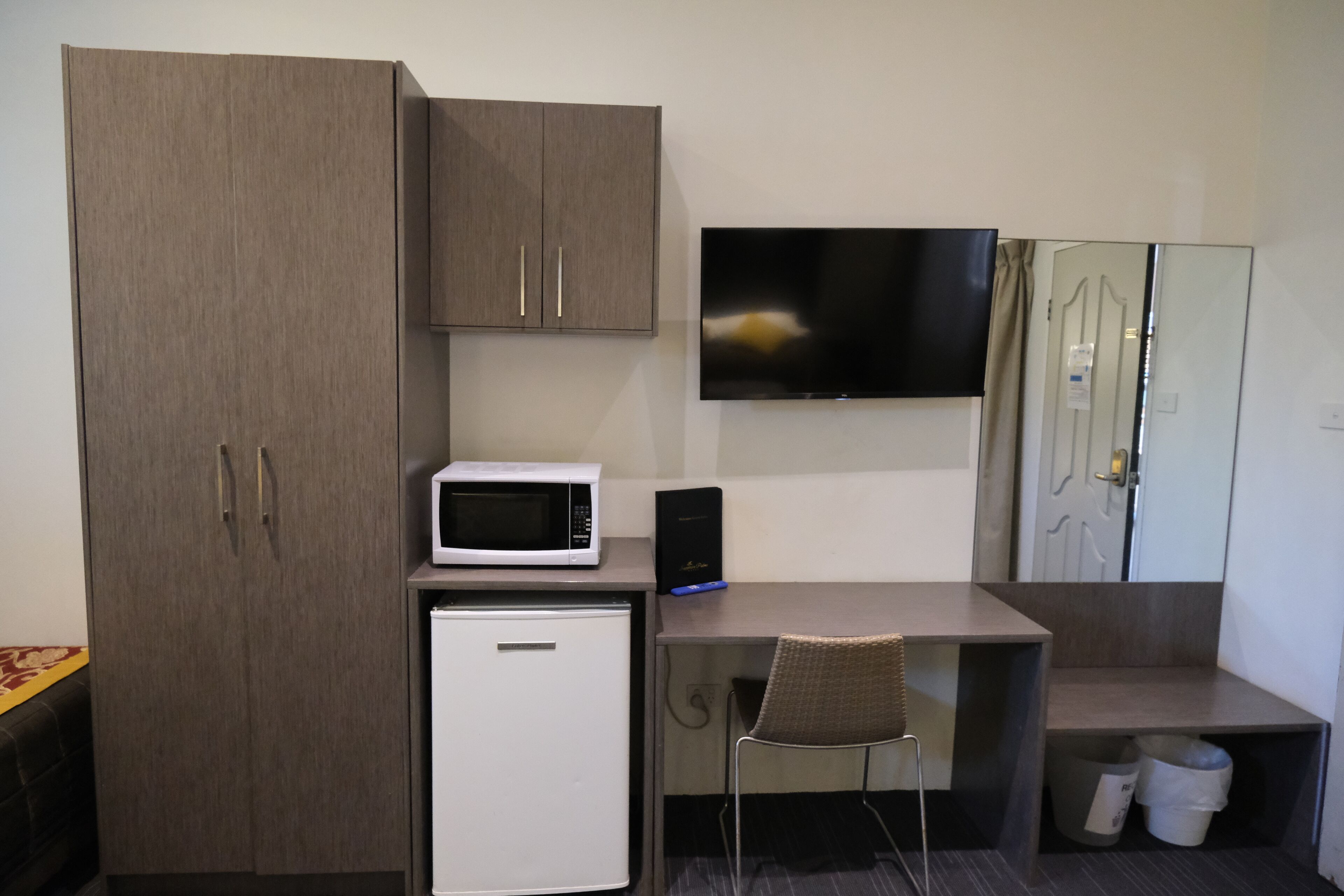 A compact room with a kitchenette, including a microwave, refrigerator, and a small cabinet. A flat-screen TV is mounted on the wall, and a chair is placed in front of a desk.