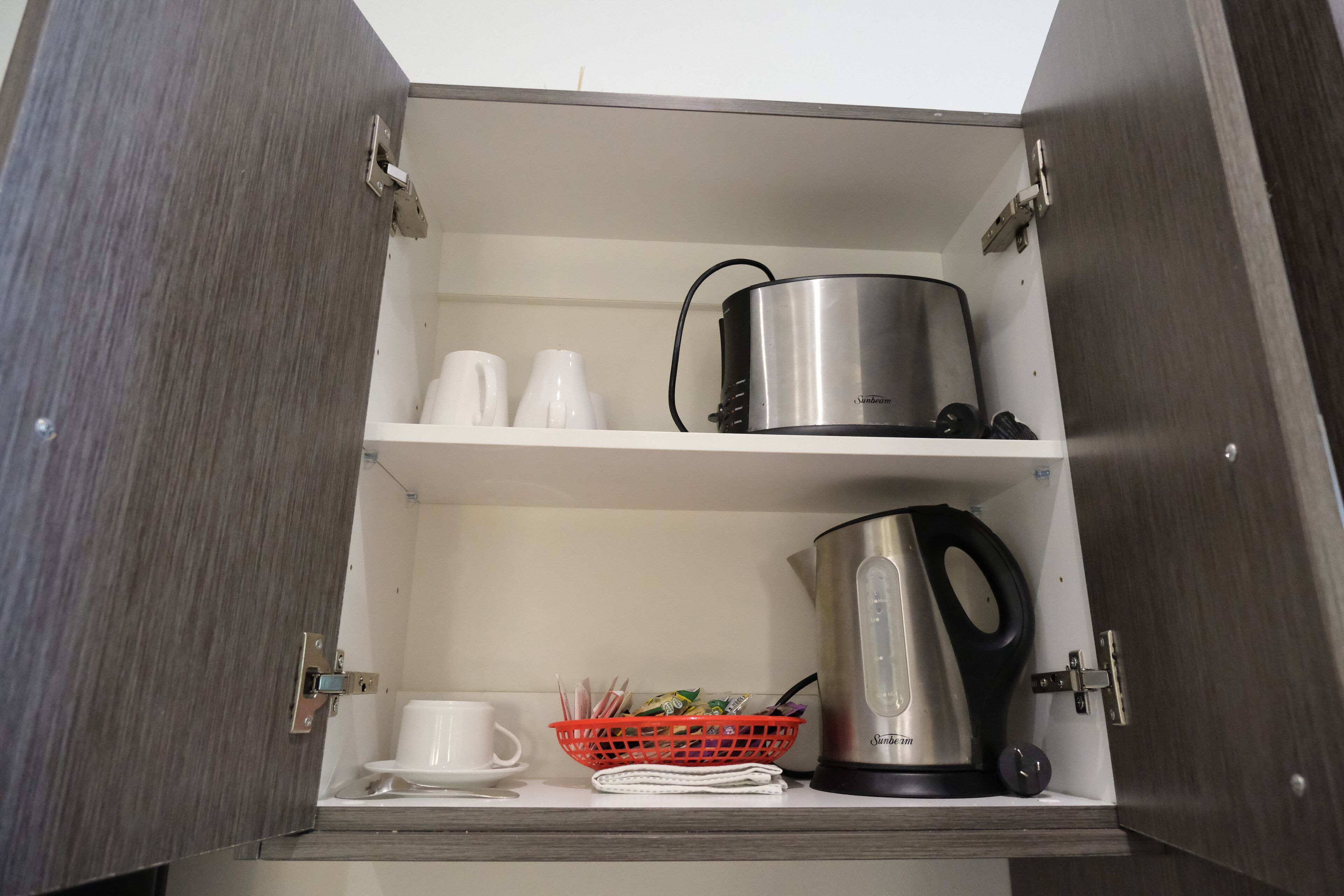 Un meuble de cuisine contenant une casserole en acier inoxydable, une bouilloire, un panier rouge et des tasses blanches.