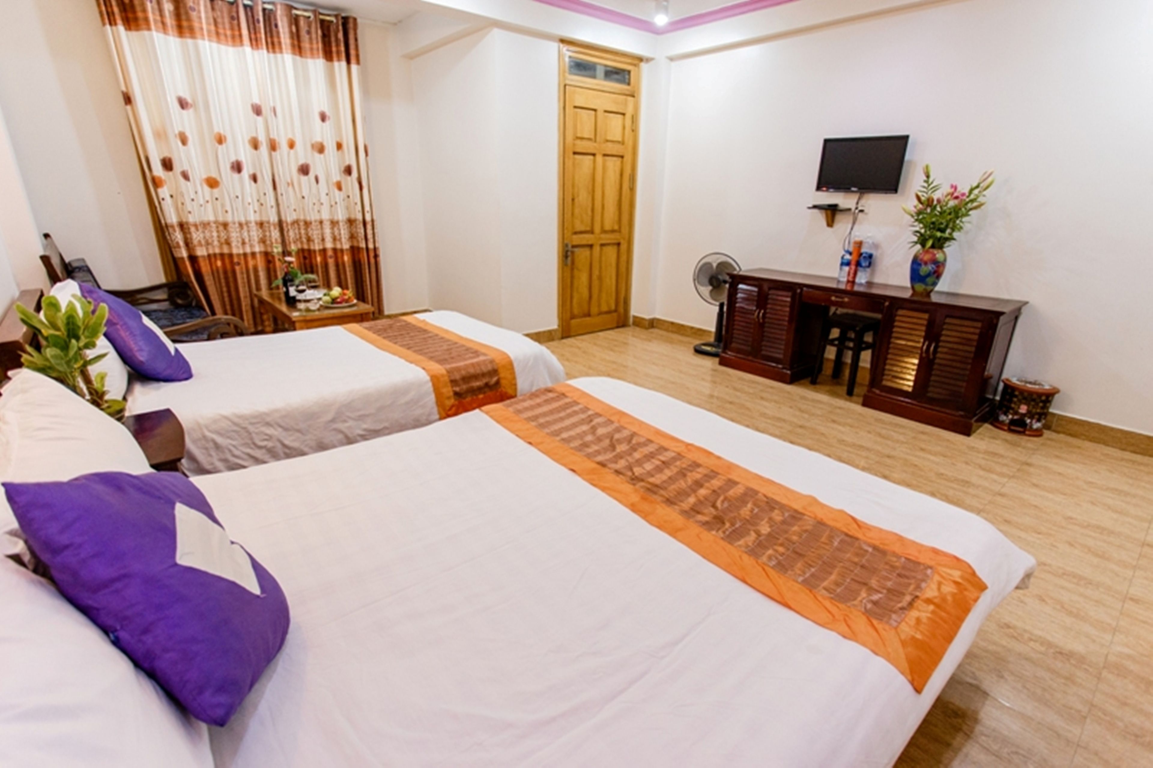 A hotel room with two beds, a wooden floor, a TV, a desk with a fan, and a door.