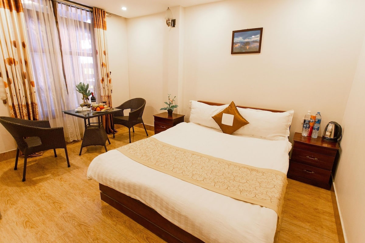 Habitación de hotel con una cama grande, un escritorio con silla, una mesita con planta y una mesita de noche con lámpara.