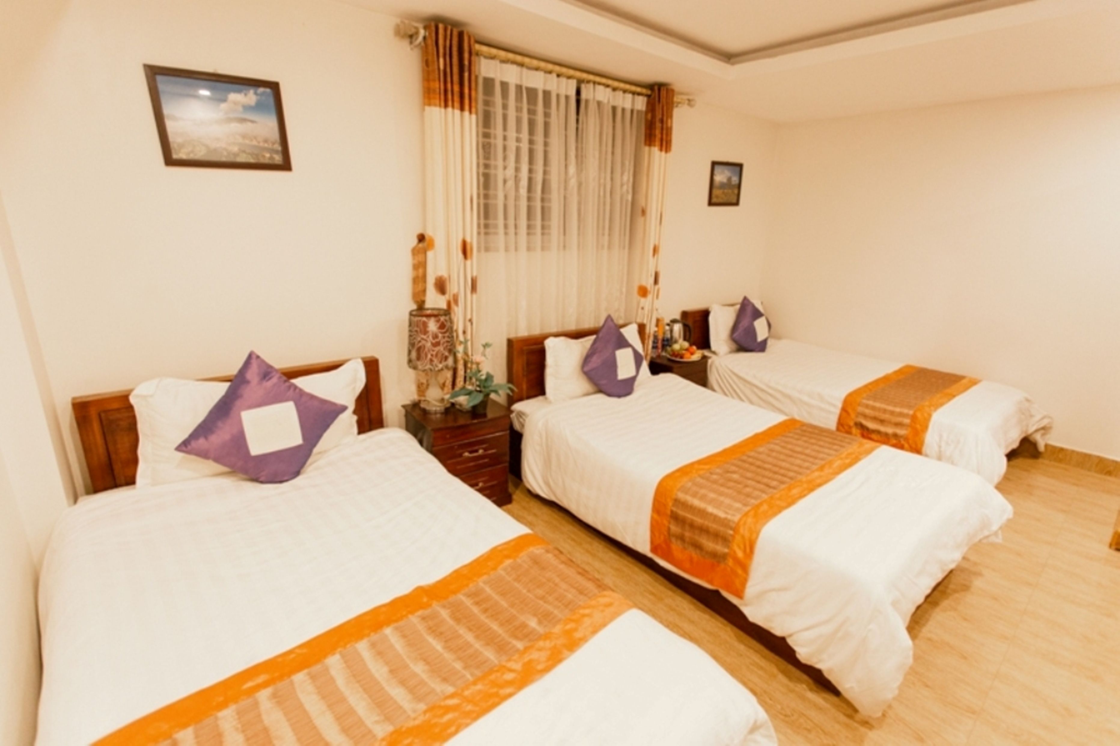 A hotel room with three single beds, each with a white bedspread and orange accents. There is a window with sheer curtains and a picture frame on the wall.