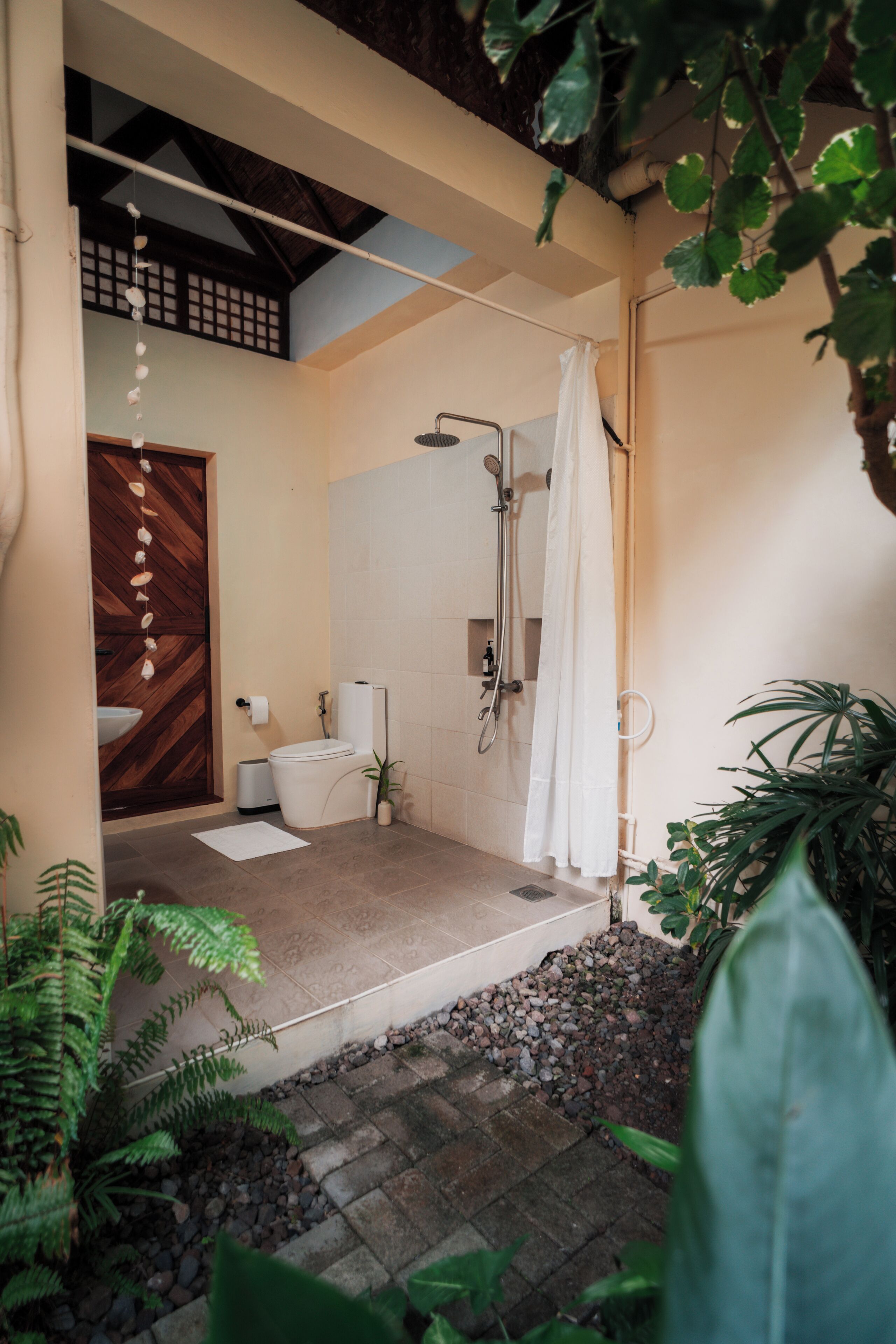 Une salle de bain avec douche, toilettes et lavabo, entourée de plantes et bordée d’un chemin en pierre.