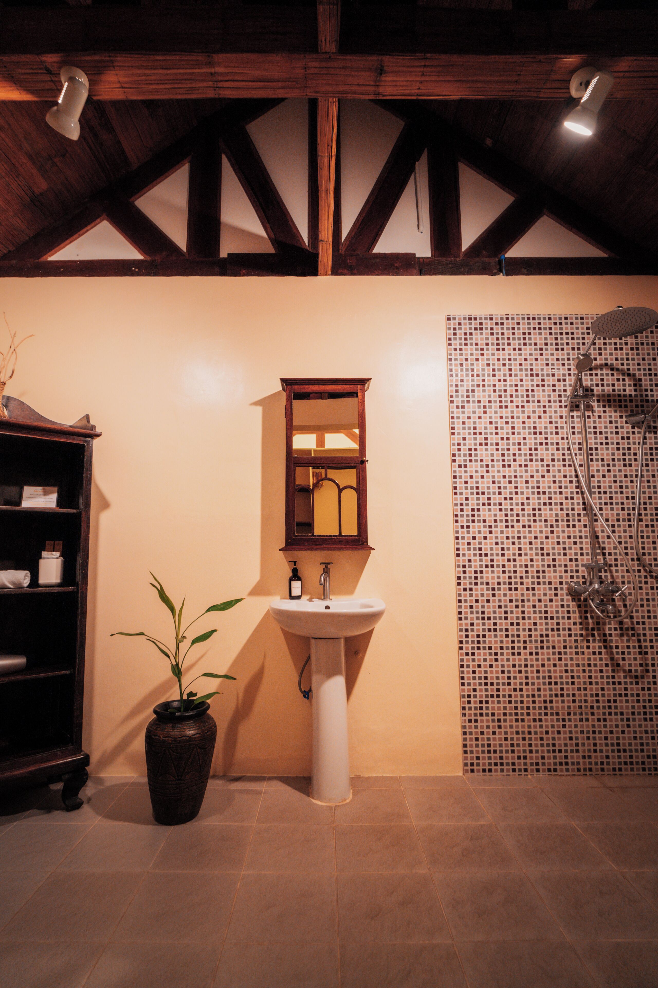 A bathroom with a wooden ceiling, a sink, a mirror, a shower, and a potted plant.