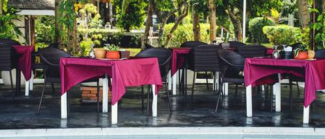 Zona junto a la piscina con mesas cubiertas con manteles rojos, sillas de mimbre y una estructura con techo de paja al fondo.