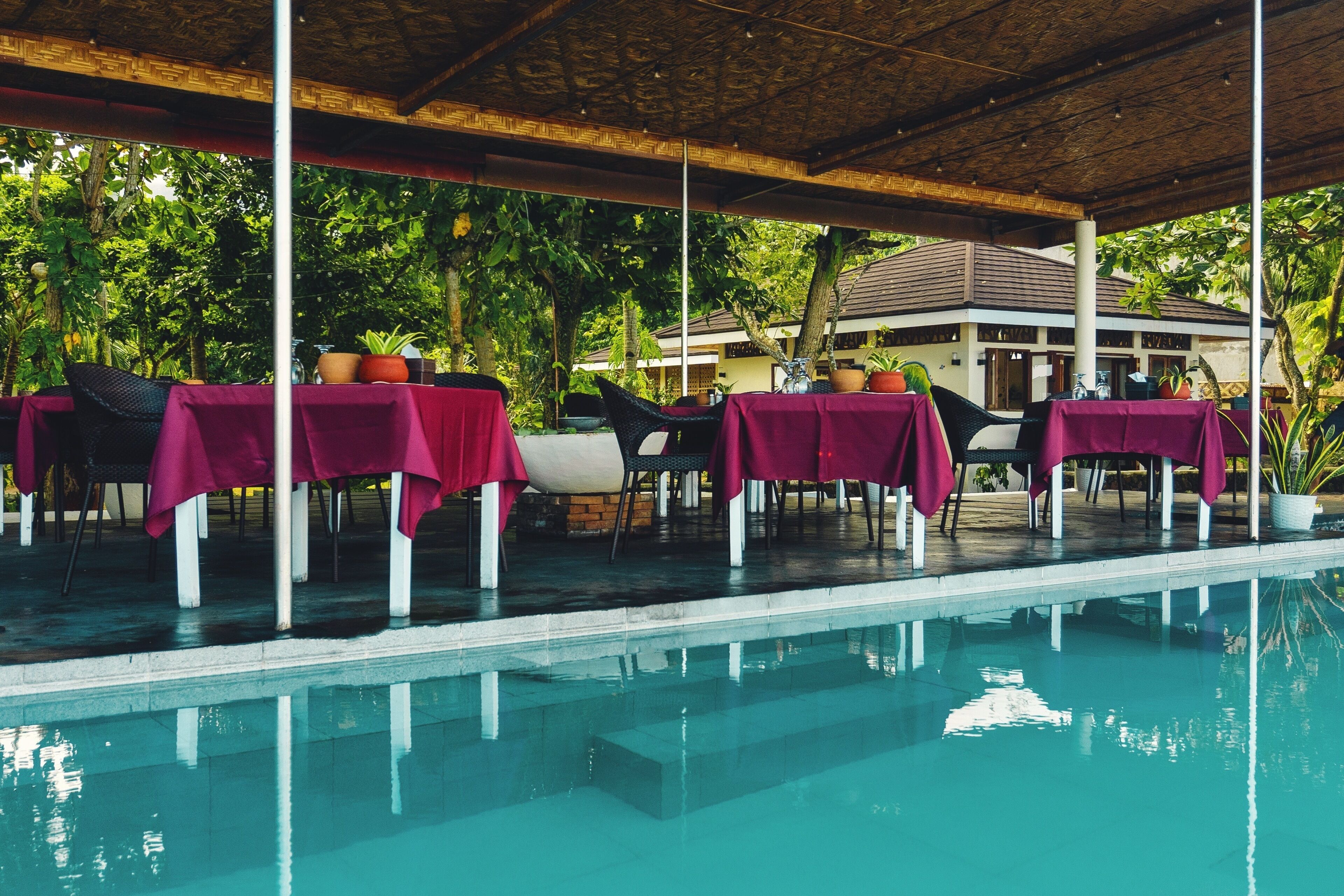 An outdoor pool area with tables covered in red tablecloths, surrounded by greenery and a building in the background.