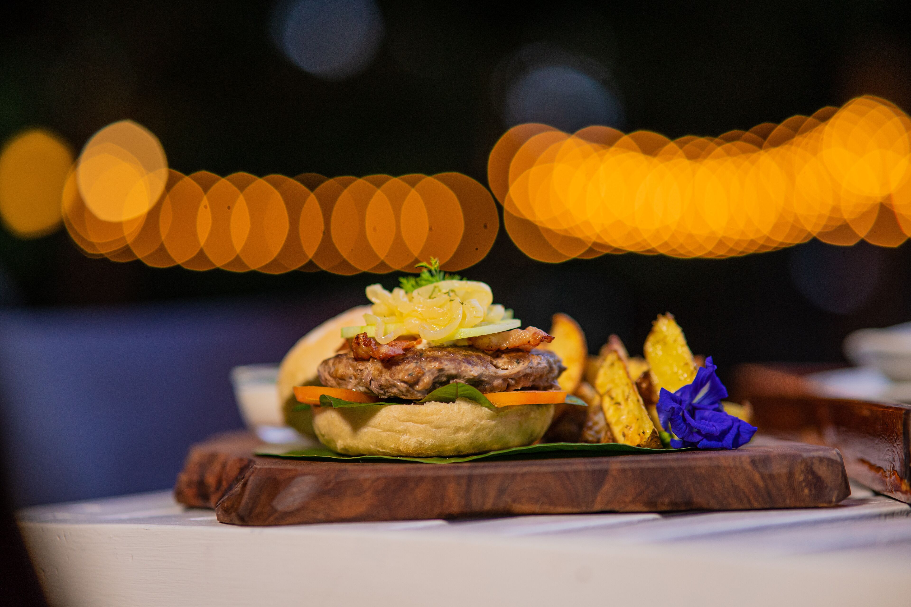 Ein Burger mit Speck, Salat und Tomate auf einem Brötchen, serviert mit Pommes und einer Blume auf einer Holzplatte.