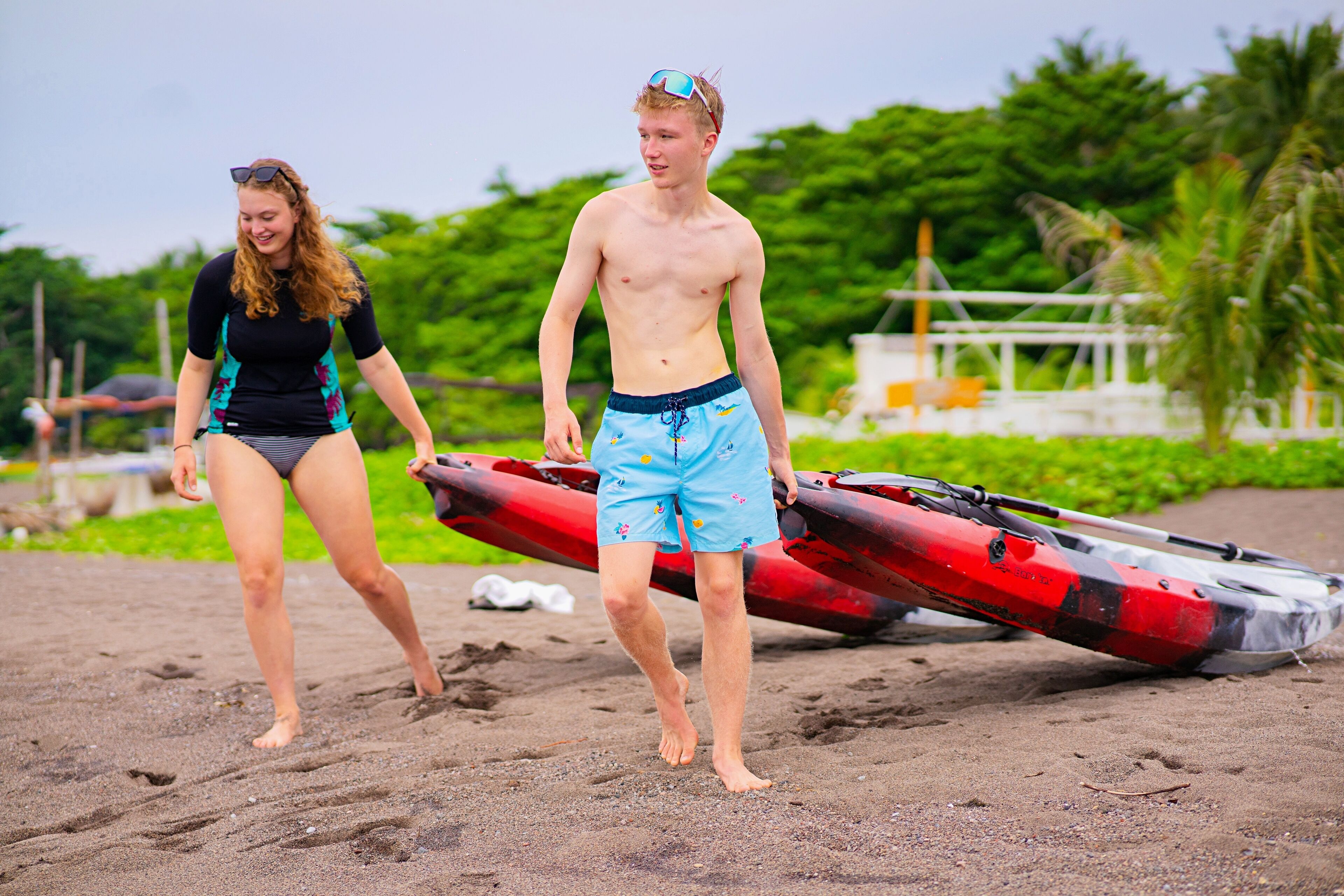 Two people walking on a sandy beach with a red kayak and a white boat in the background.
