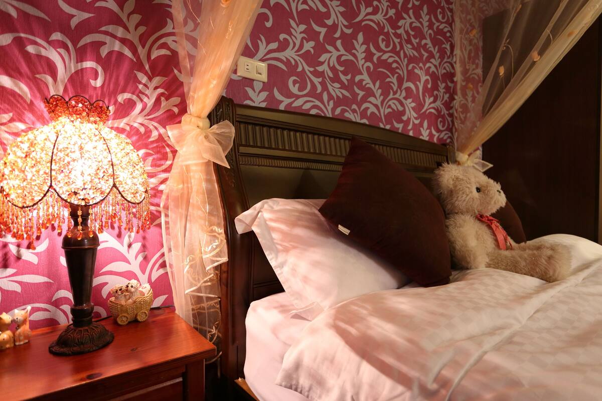 A neatly made bed with a teddy bear, a decorative lamp, and a patterned wall.