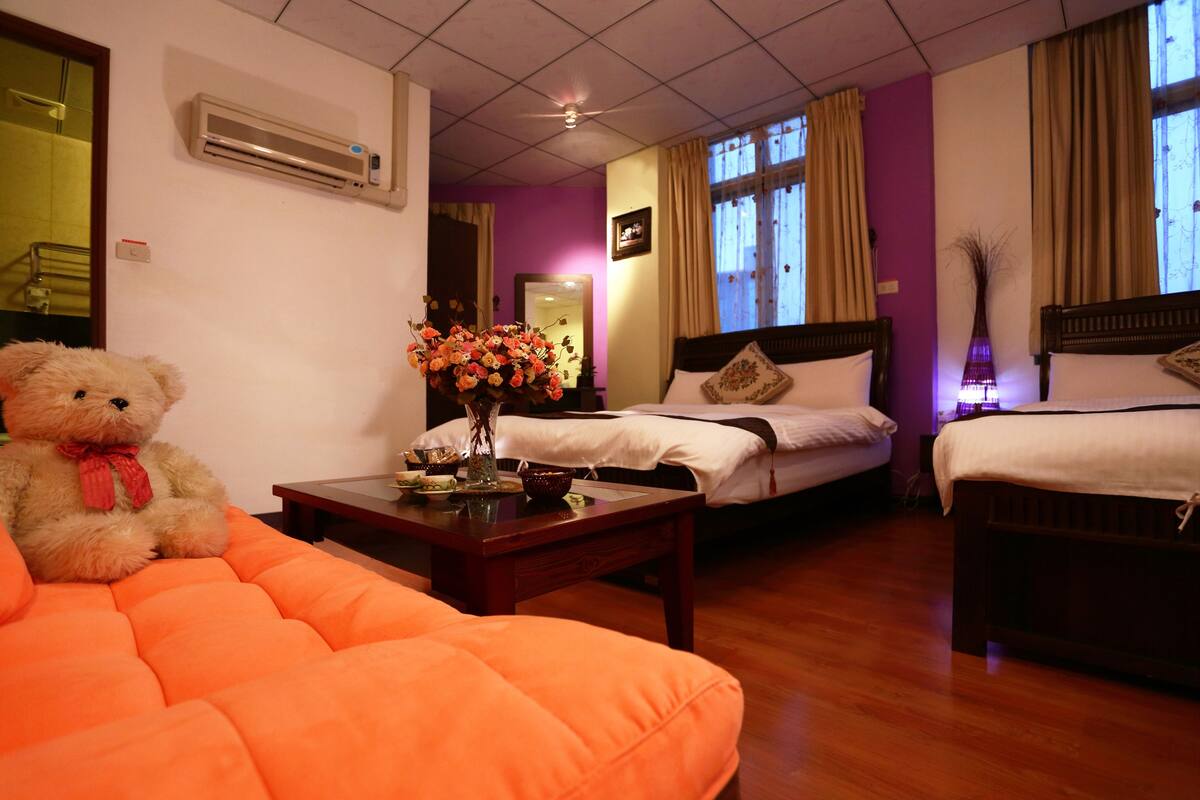 A hotel room with two beds, a sofa, a table with a vase of flowers, and an air conditioning unit on the wall.