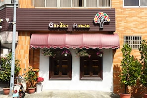 A building with a sign reading "Garden House" and a tree logo. There are two windows with brown frames and a red awning.